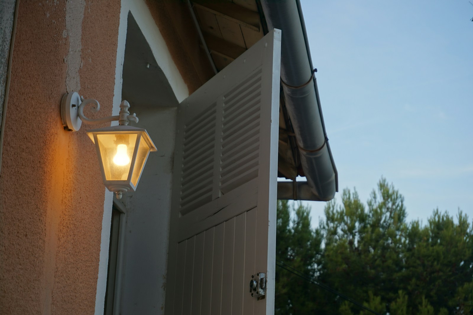 Outdoor lamp illuminates open white shutters on building.
